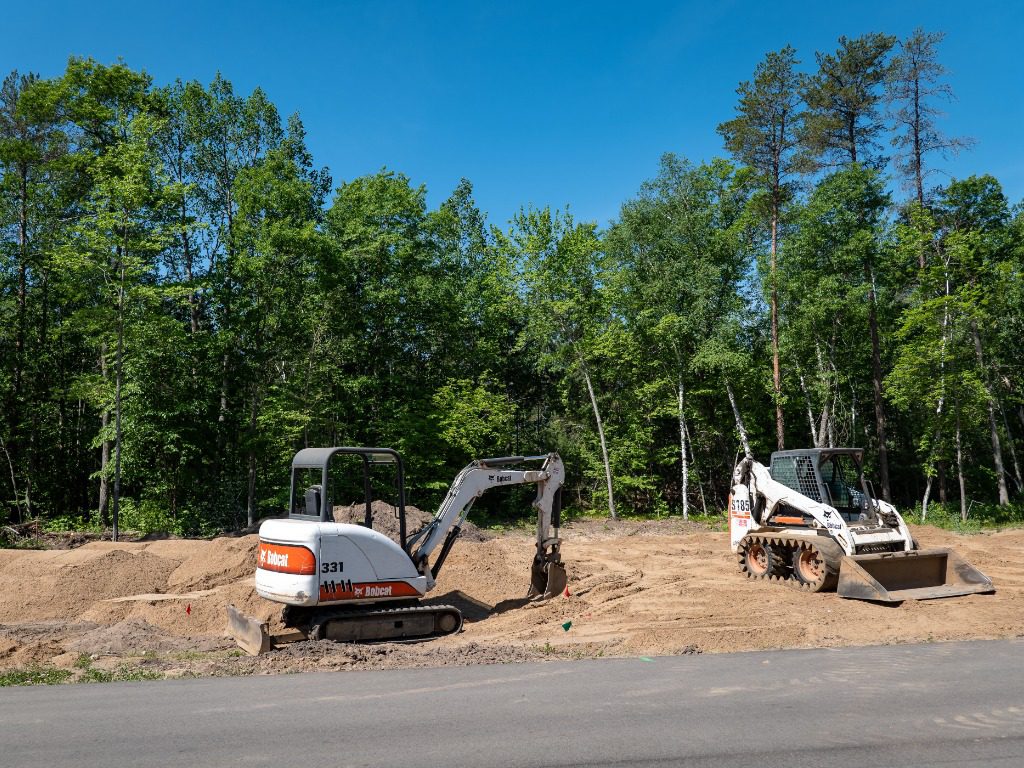 CROW WING CO, MN - 6 JUN 2021: A Bobcat mini excavator and front loader, with skid steer tracks, sits on dirt at a tree lined empty building lot, in preparation for new home construction.
